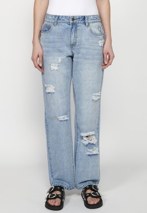 Jean en denim bleu clair avec une coupe décontractée, présentant des zones déchirées et des ourlets effilochés. Associé à des sandales noires ornées de quincaillerie décorative.