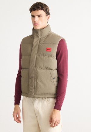 Young man wearing a beige quilted vest with a red logo patch over a long-sleeve burgundy shirt and light-colored pants, standing against white background.