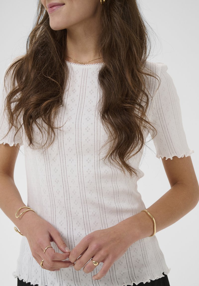 Femme aux longs cheveux bruns ondulés portant un haut blanc à manches courtes avec motifs et des bijoux en or, les mains jointes devant la taille.