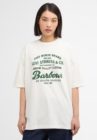 Kreemivärvi ülekaaluline T-särk, mis on valmistatud puuvillast ja millel on roheline trükisteksti disain. Lühikesed varrukad ja ümar kaelus.