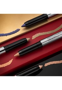 Trois crayons à yeux avec des tubes noirs : l'un doré, l'un bronze et l'un violet. Échantillons brillants sur des arrière-plans rouges et dorés.