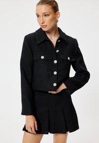 Zwart tweed cropped jasje met dubbele borstzakken, versierd met parelachtige knopen; gecombineerd met een bijpassende geplooide minirok.