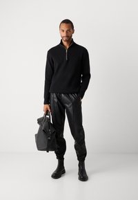 Hombre vestido con un suéter negro con cremallera, pantalones jogger de cuero negro, botas negras, sosteniendo un gran bolso de cuero negro en un estudio blanco.