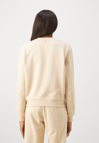 Felpa beige con collo rotondo e polsini a coste, caratterizzata da un piccolo dettaglio circolare sul retro. Tessuto morbido e liscio con una vestibilità rilassata.