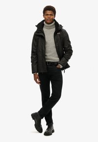 Sort vandtæt jakke med høj krave, grå sweater under, sorte bukser og sorte støvler. Modellen står i en neutral positur.