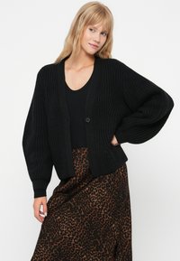 Mustapintainen neulecardigan, jossa on rento leikkaus, ylisuuret hihat ja yksi nappi kiinnityksessä, yhdistetty leopardikuvioiseen hameeseen.