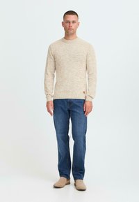 Man staat, draagt een beige gebreide trui, blauwe spijkerbroek en beige suède laarzen, tegen een effen lichte achtergrond.
