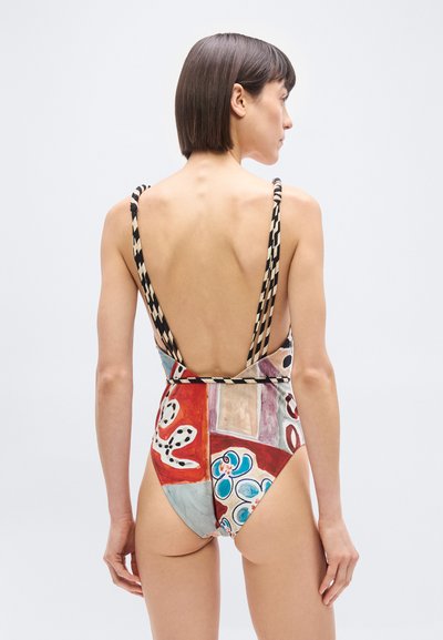 Γυναίκα με κοντά σκουρόχρωμα μαλλιά, που φοράει ολόσωμο μαγιό πολύχρωμο με λουριά με σχέδια, γυρισμένη πλάτη σε απλό φόντο.