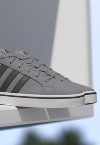 Zapatillas Adidas grises con rayas negras y suelas blancas, exhibidas sobre una superficie blanca con un fondo claro.