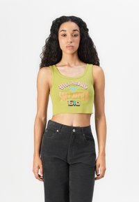 Young woman with long curly hair wearing a green cropped tank top with "Billabong Surf Africa 1640" text and black high-waisted pants.