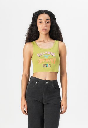 Young woman with long curly hair wearing a green cropped tank top with "Billabong Surf Africa 1640" text and black high-waisted pants.
