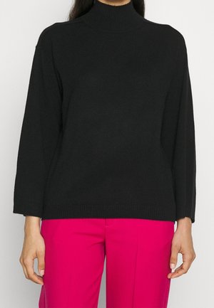 Personne portant un pull à col roulé noir à manches longues et un pantalon rose vif, debout devant un fond uni.