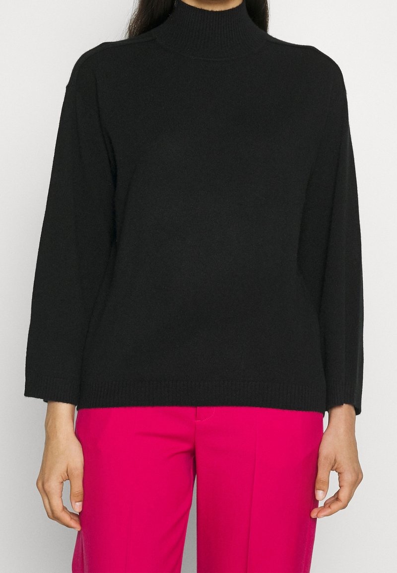 Personne portant un pull à col roulé noir à manches longues et un pantalon rose vif, debout devant un fond uni.