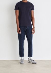 Man som står klädd i en marinblå t-shirt, mörkblå jeans, vita strumpor och grå sneakers mot en plain vit bakgrund.