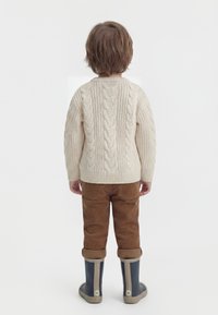 Kind mit kurzen braunen Haaren, das einen cremefarbenen Pullover mit Zopfmuster, braune Kordhosen und marineblaue Regenstiefel trägt und mit dem Rücken zur Kamera steht.