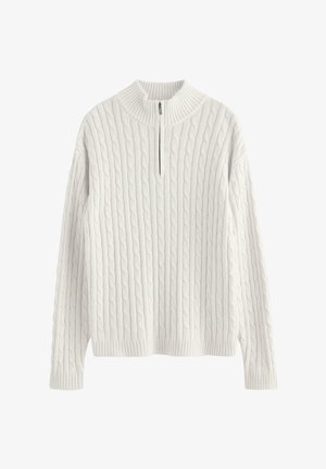 Jongen staande en glimlachend, gekleed in een witte kabelgebreide trui met rits, lichtblauwe spijkerbroek en witte sneakers, tegen een effen witte achtergrond.