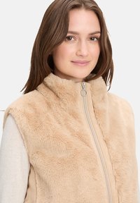 Beżowa kamizelka z futra syntetycznego z wysokim kołnierzem, pełnym zamkiem i gładką podszewką. Miękka tekstura i subtelne różnice w wzorze futra.