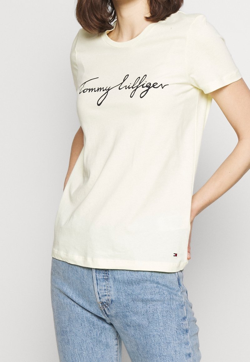 T-shirt à manches courtes jaune clair en coton, avec un logo en cursive noir "Tommy Hilfiger" sur le devant et un petit logo sur l'ourlet.