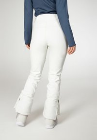 Witte skibroek met een uitlopende snit, voorzien van ritsen bij de pijpen en een gladde, glanzende textuur. Gecombineerd met lichtgrijze sportschoenen.