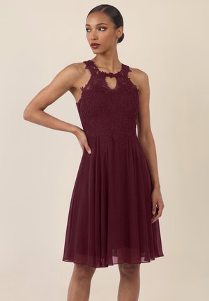 Frau trägt ärmelloses knieumspielendes Kleid aus burgunderroter Spitze und Chiffon, Hand in der Hüfte, blickt nach links vor beigem Hintergrund.