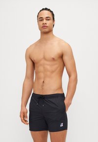 Svarte badeshorts med snøring i livet og logo-lapp. Glatt, lett materiale; kne-lengde design; avslappet, sporty utseende.