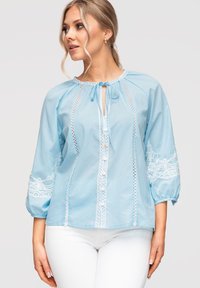Femme portant un chemisier bleu clair avec des détails en dentelle blanche, manches ballon trois-quarts et pantalon blanc, regardant sur le côté devant un fond uni.