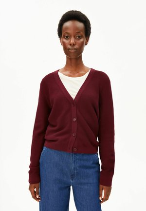 Frau, die eine burgunderrote, geknöpfte Strickjacke über einem weißen Top und blauen Jeans trägt und vor einem schlichten weißen Hintergrund steht.