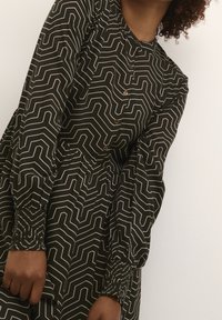 Robe verte avec un motif géométrique blanc, manches longues avec des poignets smockés, et une fermeture à boutons sur le devant. Tissu lisse avec une coupe décontractée.