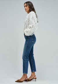 Mujer con blusa blanca con volantes, jeans azul de cintura alta y tacones marrones tipo slingback, de pie contra un fondo claro y sencillo.
