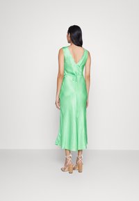 Satin grünes Kleid mit gewelltem Ausschnitt, offener Rückseite und Knoten in der Taille; kombiniert mit weißen Riemchen-Pumps und Kork-Plateausohle.