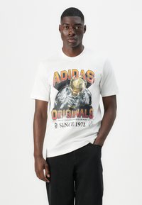 Hvid bomulds t-shirt med et iøjnefaldende sort og orange "ADIDAS ORIGINALS" grafik med en trofæ- og jorddesign, afslappet pasform og korte ærmer.
