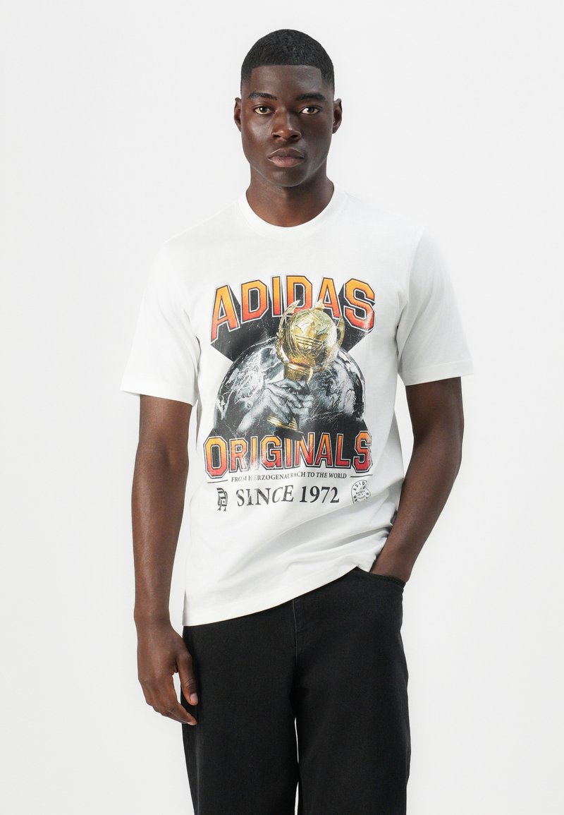 Camiseta de algodón blanca con un gráfico audaz en negro y naranja de "ADIDAS ORIGINALS" que incluye un trofeo y un diseño de la Tierra, corte relajado y mangas cortas.