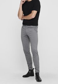 Graue Hose mit schmaler Passform, gefertigt aus einem glatten Stoff. Verfügt über Vordertaschen, Gürtelschlaufen und ein dezentes strukturiertes Muster. Trägt ein schwarzes T-Shirt dazu.