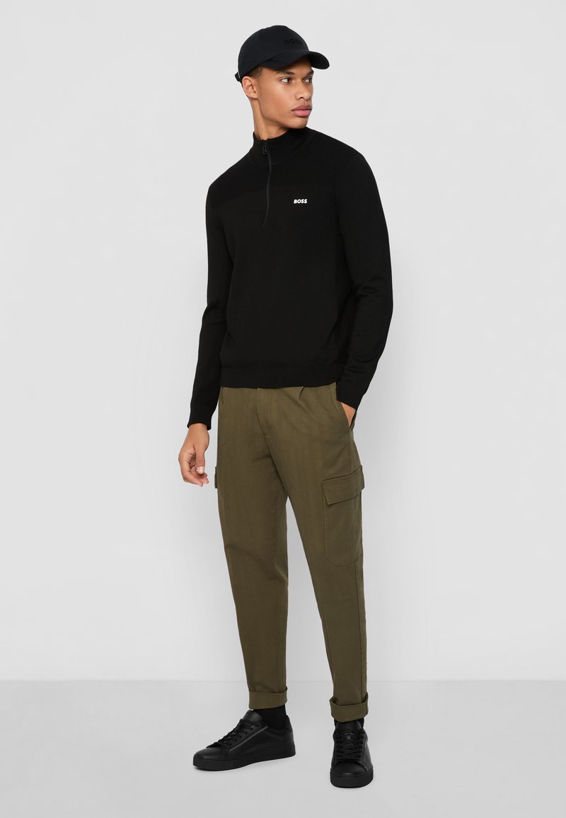 Schwarzer Zip-Kragen-Pullover mit kleinem weißen Logo, kombiniert mit olivfarbenen Cargo-Hosen mit Seitentaschen und schwarzen Sneakern.