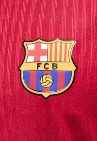 Το έμβλημα της FCB με κόκκινο σταυρό, κίτρινες και κόκκινες ρίγες και μπλε και κόκκινες κάθετες ρίγες σε υφασμένο κόκκινο φόντο.