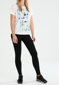 Camiseta gráfica blanca con letras azules y verdes "EMPEZAR AHORA," combinada con leggings negros y zapatillas deportivas negras. Textura suave, corte relajado.