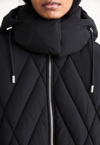 Άτομο που φοράει μαύρο καπιτονέ μπουφάν με μεγάλο επενδυμένο γιακά και ορατό ασημένιο φερμουάρ, κοντινή λήψη στην περιοχή του λαιμού και του στήθους.