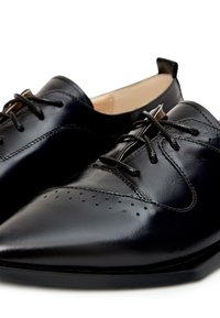 Brogues en cuir noir avec un bout pointu, présentant des perforations décoratives, un design minimaliste élégant et des lacets noirs sur un intérieur beige.