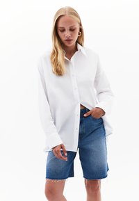 Camicia bianca a maniche lunghe con colletto e chiusura a bottoni abbinata a pantaloni shorts in denim blu con orli sfrangiati, creando un look dal fit rilassato.
