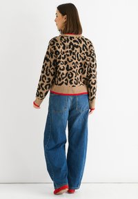 Leopardenmuster-Pullover in Taupe und Schwarz mit roten Akzenten; lockere Passform, gerippte Bündchen und Saum. Kombiniert mit hoch taillierten blauen Jeans.