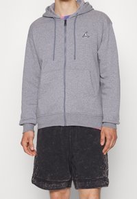 Felpa grigia con zip realizzata in un tessuto misto cotone, con tasca a marsupio e logo Jumpman nero sul lato sinistro del petto. Vestibilità ampia.