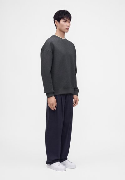 Jeune homme debout portant un sweat-shirt gris foncé, un pantalon noir ample et des baskets blanches sur un fond clair uni.