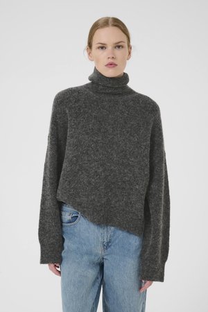 Mørkegrå genser i ullblanding med høy hals, vide ermer og asymmetrisk kant, kombinert med lys blå jeans.