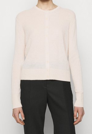 Personne portant un cardigan boutonné rose clair à manches longues et un pantalon taille haute noir, debout devant un fond uni.