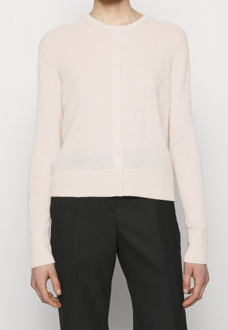 Personne portant un cardigan boutonné rose clair à manches longues et un pantalon taille haute noir, debout devant un fond uni.