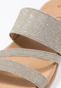 Sandales argentées pailletées avec des brides larges texturées et une semelle intérieure beige douce. Présentent une palette de couleurs neutres et une finition lisse.