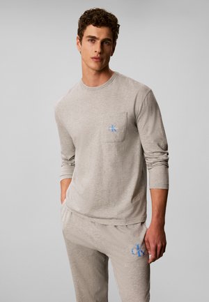 Jeune homme portant une chemise beige à manches longues et un pantalon assorti avec des logos Calvin Klein bleus sur la poche poitrine et la cuisse, debout devant un fond uni.