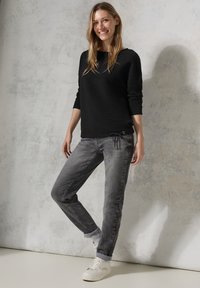 Schwarzer strukturierter Pullover mit langen Ärmeln, kombiniert mit grauen Jeans und weißen Sneakern, vor einem hellen strukturierten Hintergrund stehend.