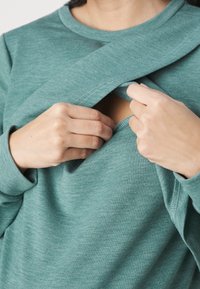 Tealfärgad långärmad topp med en överlappande design fram, som avslöjar en liten öppning under, gjord av mjukt, texturerat tyg.