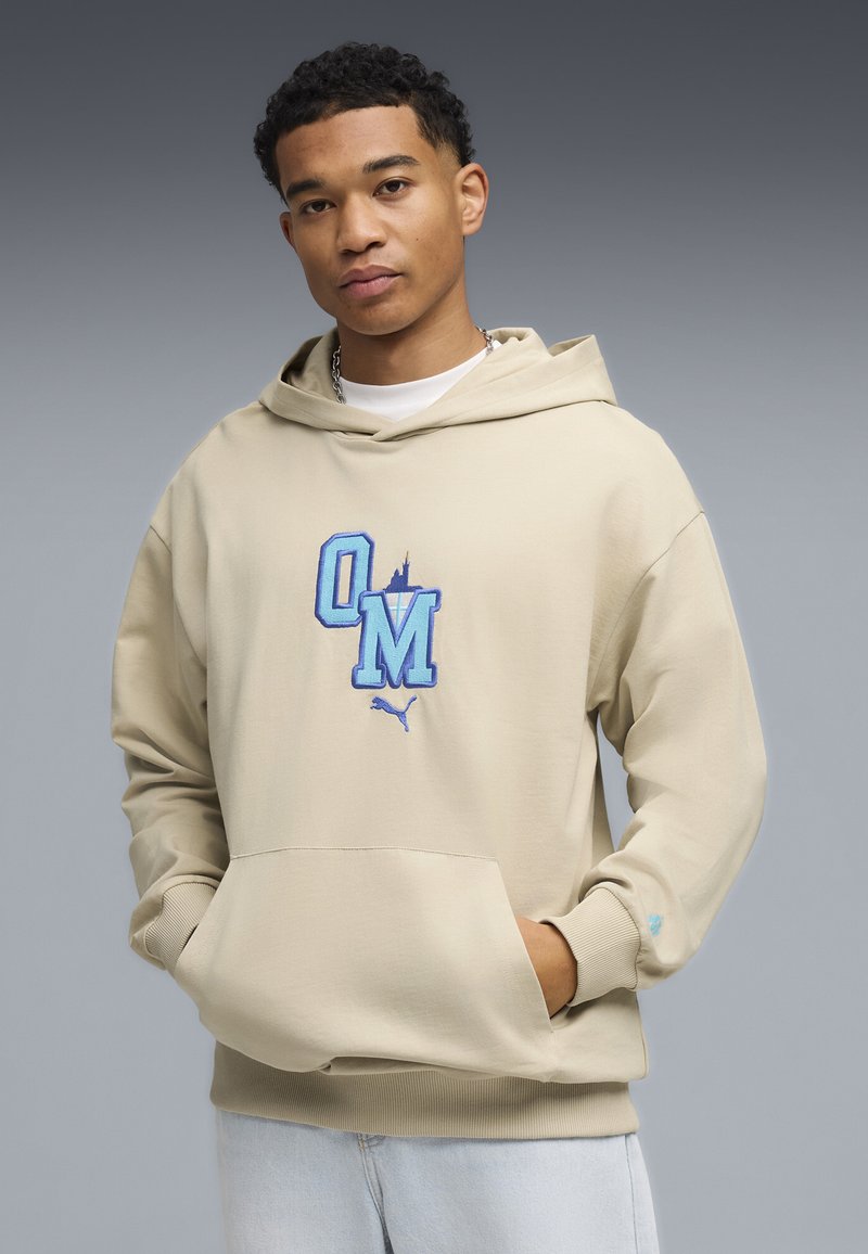 Beige Kapuzenpullover mit Kängurutasche, blau gesticktem "OM"-Logo, gerippten Bündchen und Saum. Aus weichem Stoff, legerer Schnitt.